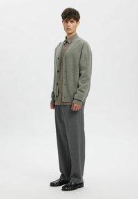 Pelēks kardigans ar pogām, kombinēts ar bēša krāsas rūtainu kreklu, pelēkām piegrieztām biksēm un melniem apaviem, attēlots uz baltas fona.