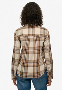 Chemise en flanelle beige avec un motif à carreaux marron et bleu marine. Présente des manches longues, un col pointu et une ourlet arrondi. Texture douce.