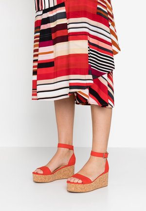 Jambes en sandales compensées rouges avec brides à la cheville, associées à une jupe midi rayée multicolore avec un ourlet asymétrique.