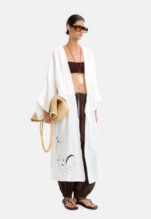 Kobieta w oversize'owym białym kimono z wycięciami, brązowym topie na ramiączkach i luźnych spodniach, trzymająca plecioną torbę i nosząca duże okulary przeciwsłoneczne.