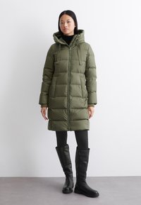 Olivengroene pufferjas met een capuchon, ritssluiting aan de voorkant en elastische manchetten. Gecombineerd met zwarte lange laarzen en leggings, wat een gestroomlijnd silhouet benadrukt.