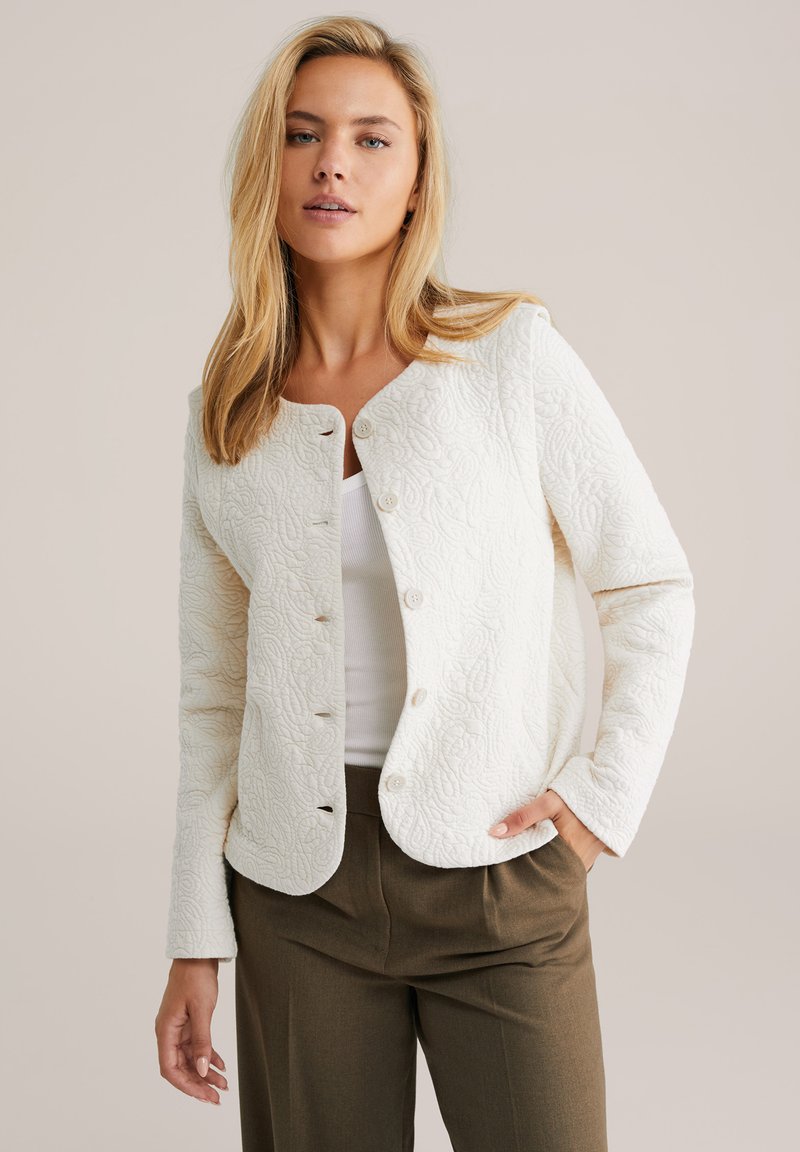 Textured white jacket with a round neckline and five button front closure. Features long sleeves and floral embossed pattern.