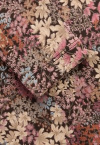 Tkanina kwiatowa z różowymi, beżowymi i niebieskimi wzorami na brązowym tle, z zbliżeniem mankietu ukazującym detale zszycia.