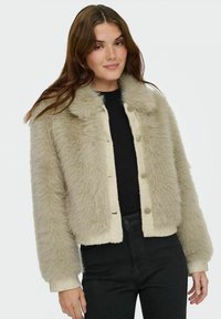 Beige faux fur jacket with a cropped design, featuring a button front and a soft, textured finish. Worn over a black top and dark jeans.