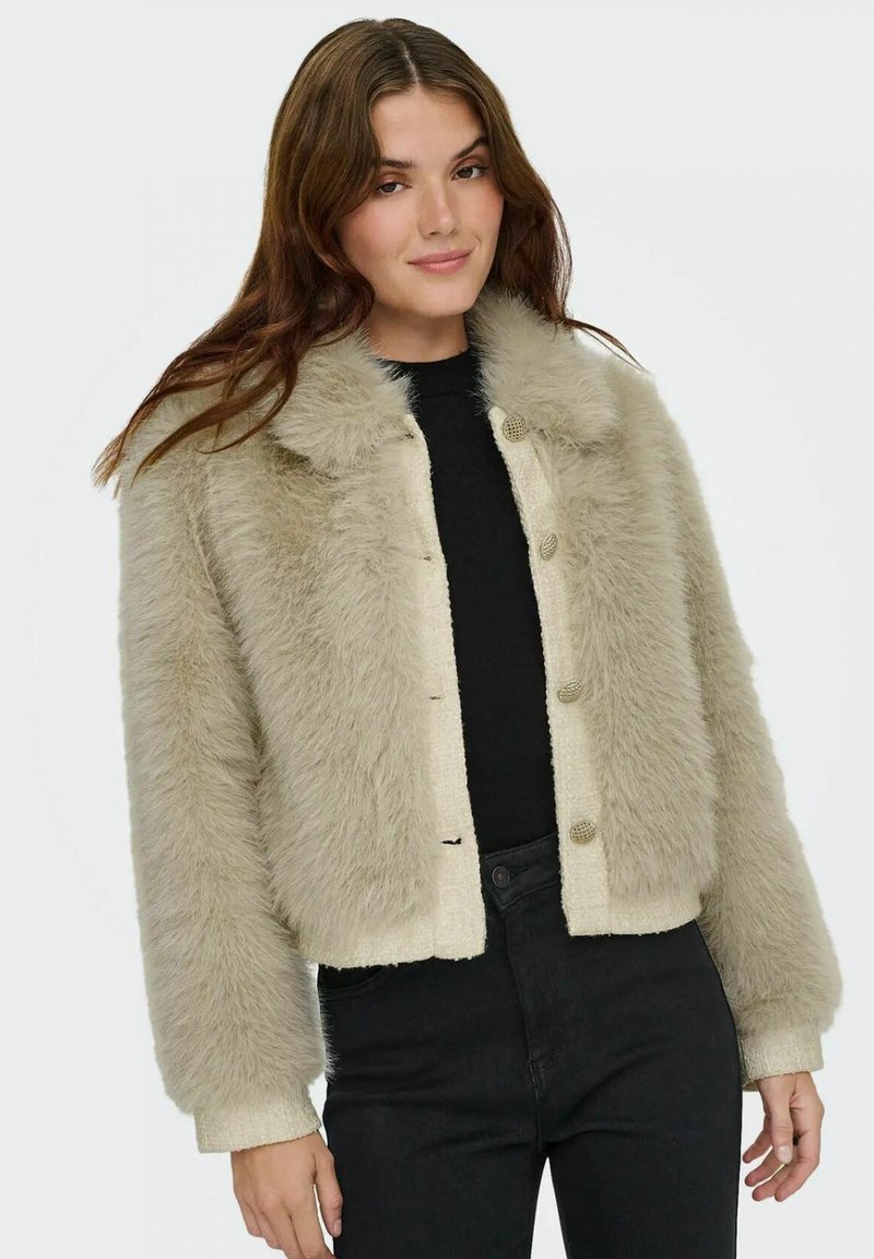 Beige faux fur jacket with a cropped design, featuring a button front and a soft, textured finish. Worn over a black top and dark jeans.