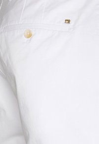 Pantalon blanc en coton avec une texture lisse, une poche avant ornée d'un accent doré subtil, et une fermeture par bouton.