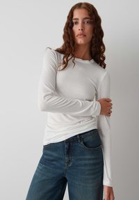 Langärmliges weißes Baumwollhemd mit Rundhalsausschnitt, kombiniert mit dunkelblauen Jeans. Hat eine figurbetonte Silhouette und eine glatte Textur.