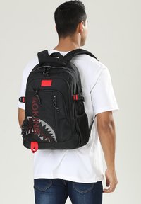 Sac à dos noir avec des accents rouges et un design de requin, comportant plusieurs compartiments zippés, des sangles réglables et un tissu texturé.