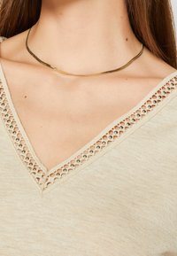 Cou au féminin portant une fine chaîne en or et un haut beige à col en V avec une bordure ornée de clous, ressemblant à de la dentelle.