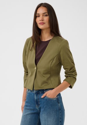 Kvinde med langt mørkt hår iført en olivengrøn kort blazer over en mørk top og blå jeans, stående med den ene hånd i lommen.
