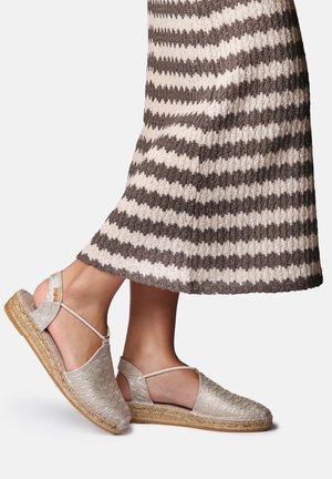 Pies con sandalias alpargatas beige tejidas con tiras en el tobillo, combinadas con una falda de punto a rayas marrón y blanca.