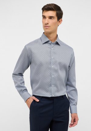 Camicia a fantasia blu con design a bottoni, caratterizzata da piccole forme geometriche, maniche lunghe e colletto classico. Abbinata a pantaloni scuri.