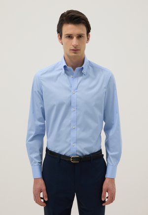 Homme portant une chemise habillée bleu clair boutonnée, rentrée dans un pantalon foncé avec une ceinture noire, debout devant un fond clair uni.