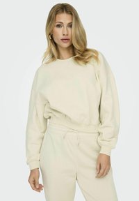 Sweatshirt color crema, corto, con colletto rotondo, maniche ampie e polsini a coste. Abbinato a pantaloni da tuta a gamba stretta coordinati.