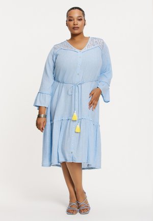 Blaues Chambray-Kleid mit Knopfleiste, Rüschensäumen und gebundenem Taillenbund. Es hat dekorative Quasten in Gelb und Weiß.