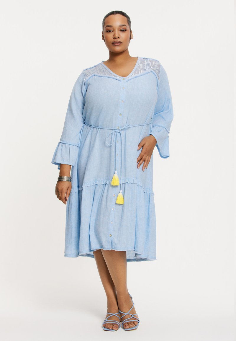Blaues Chambray-Kleid mit Knopfleiste, Rüschensäumen und gebundenem Taillenbund. Es hat dekorative Quasten in Gelb und Weiß.