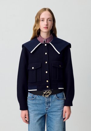 Veste cropped navy avec grand col, devant à boutons, poches à rabat et bordures blanches contrastées ; portée sur une chemise à carreaux et un jean.