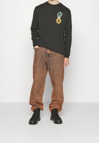 Black long-sleeve t-shirt with spiral and sun graphics, paired with loose, textured, orange-brown jeans and black ankle boots.