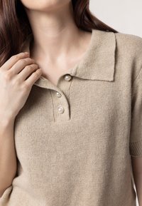 Beige gebreide polo shirt met een kraagnek, voorzien van drie parelachtige knopen en korte mouwen, weergegeven in een zachte, gestructureerde stof.
