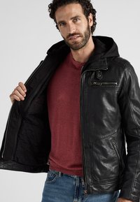 Zwarte leren jas met een capuchon, voorzien van ritsafsluitingen, voorzakken en een gevoerde binnenkant, gedragen over een rood shirt.