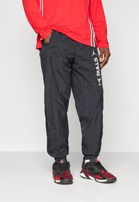 Man wearing black Chicago Bulls track pants with logo and black-red athletic shoes, standing against a plain background.