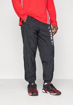 Homme portant un pantalon de survêtement noir des Chicago Bulls avec logo et des chaussures de sport noires et rouges, se tenant devant un fond uni.