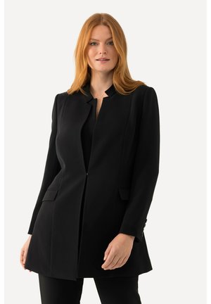 Cappotto lungo nero su misura con collo a scollo, tasche frontali e polsini con bottoni. Texture liscia e silhouette strutturata.