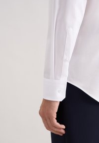 Witte overhemd met lange mouwen, voorzien van een knoopmanchet en een gladde textuur, met een lichte glans en een getailleerde pasvorm, gecombineerd met donkere broeken.