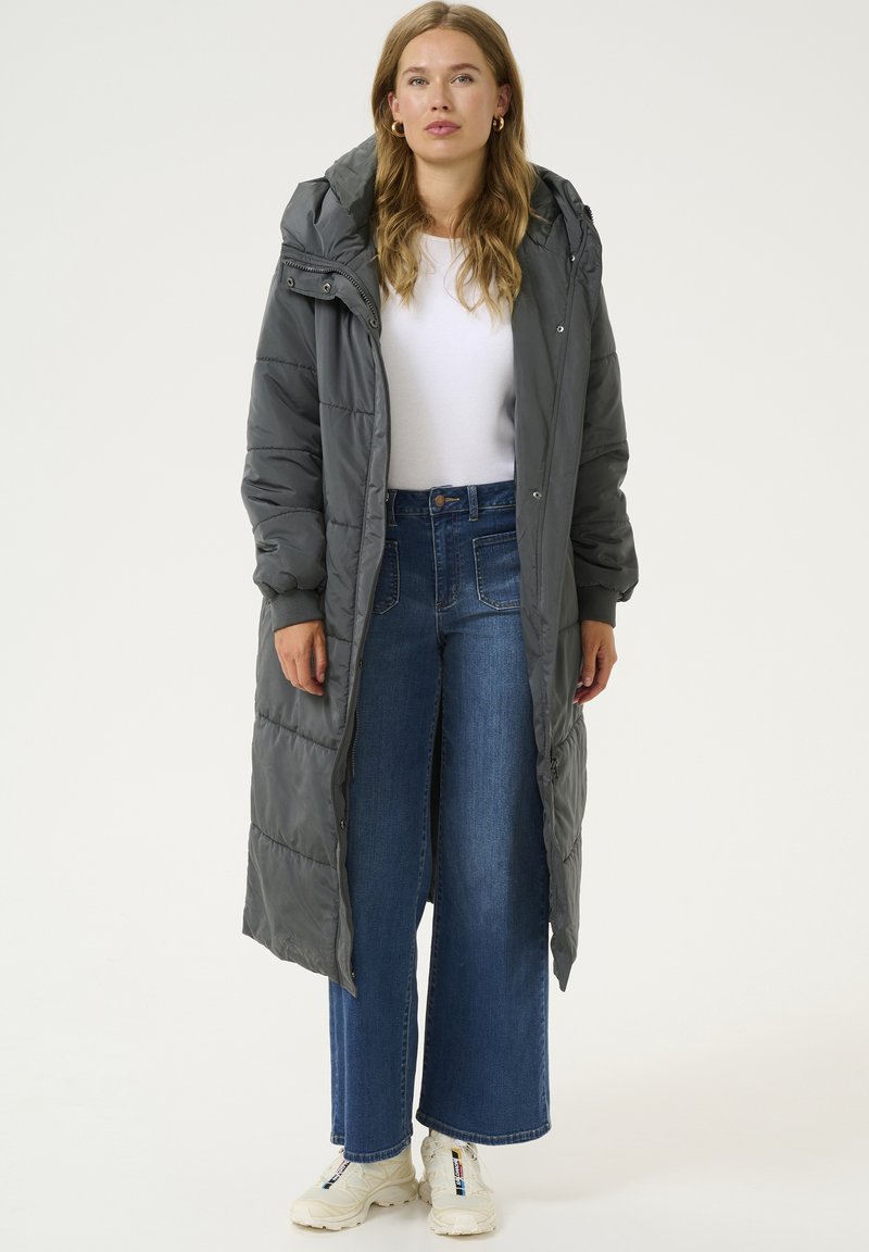 Manteau long gris rembourré avec capuche, doté d'une fermeture éclair à l'avant, de poignets côtelés et de poches latérales, assorti à un jean bleu à jambes larges et des baskets blanches.