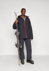 Μαύρο σκιώτικο τζάκετ με κόκκινες λεπτομέρειες, γκρι μπλούζα, σκούρες παντελόνες σκι, κρατώντας σκι με δέστρες, στέκοντας μπροστά σε ουδέτερο φόντο.