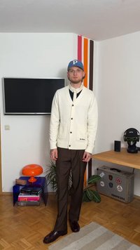 White knitted cardigan with v-neck and front pockets, paired with dark brown pants and shiny black shoes. Blue cap with logo accent.