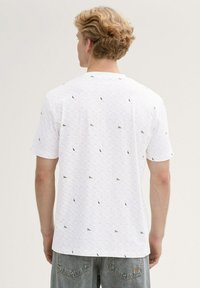 Hombre rubio con cabello corto y rizado que lleva una camiseta blanca de manga corta con estampado y jeans de color azul claro, de espaldas contra un fondo uniforme.