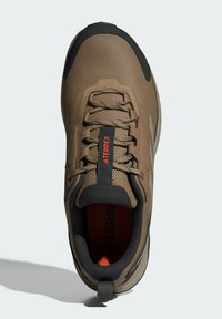 Brązowe buty sportowe z siateczkową cholewką, czarnymi akcentami i czerwonym logo. Posiadają wyściełany kołnierz, kontrastującą gumową podeszwę oraz teksturowany system sznurowania.