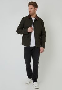 Veste vert foncé à col brun avec des boutons-pression, deux poches avant, portée sur un t-shirt blanc et associée à un jean foncé et des baskets blanches.