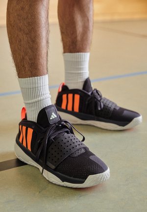 Jambes avec des chaussettes blanches montantes portant des chaussures de sport noires avec des bandes orange et des semelles blanches, debout sur le sol d’un terrain couvert.