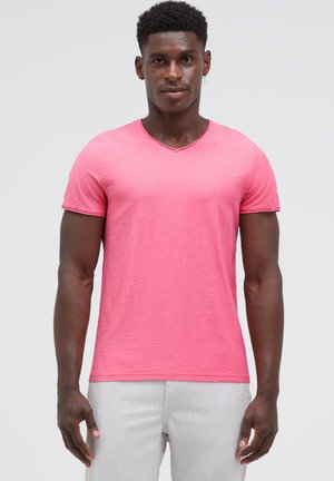Man wearing a bright pink short-sleeve V-neck shirt and light gray pants, standing against a plain light background.