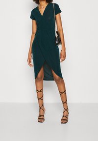 Donna che indossa un vestito avvolgente verde scuro con maniche corte, sandali neri con tacco alto e cinturini, e una piccola borsa nera con tracolla in catena dorata.