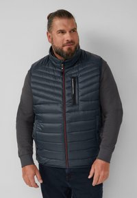 Herren Steppweste in Marineblau mit hohem Kragen, Reißverschluss-Brusttasche und horizontaler Steppung. Mit einem kontrastierenden roten Reißverschluss.
