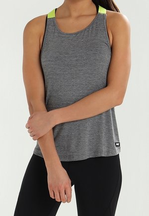 Kobieta w szarej sportowej koszulce na ramiączkach z neonowożółtymi paskami na ramionach i czarnych legginsach, stojąca ze skrzyżowanymi ramionami.