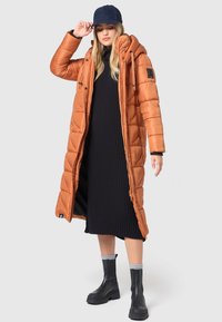 Long, puffy, orange coat with a hood, featuring a zipper and snap buttons. Worn over a black ribbed dress and paired with black boots.