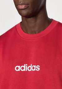 Közeli kép egy személyről, aki piros Adidas kereknyakú pólót visel, amelyen fehér betűkkel van nyomtatva a márkanév a mellkasnál.