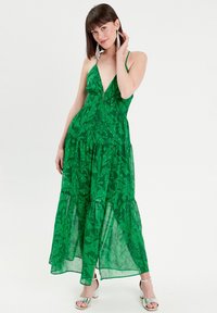 Groene maxi-jurk met een diepe V-hals, laagjes en een bladprint. Voorzien van verstelbare bandjes en een gestructureerde stof, gecombineerd met zilveren hakken.