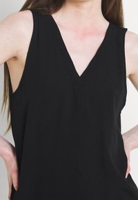 Žena v bezramenném černém topu s výstřihem do V, s prsty opřenými o boky, světlou pletí a dlouhými hnědými vlasy viditelnými na levé straně.