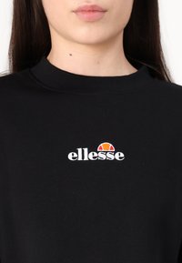 Svart hettegenser med rund hals. Har hvit brodert merkevare navn "ellesse" og en fargerik sol-logo. Myk stoff tekstur.