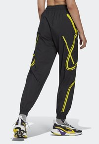 Osoba v černých teplákách s žlutými abstraktními liniemi a černo-bílo-žluto-fialových teniskách, stojící s jednou nohou zvednutou.