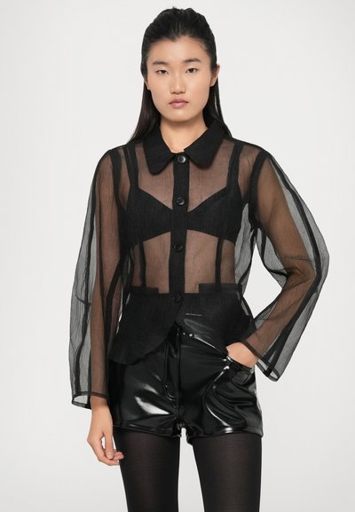 Čierna polpriesvitná blúzka s dlhými, voľne splývajúcimi rukávmi, špicatým golierom a šiestimi prednými gombíkmi; nosená cez lesklé čierne šortky.