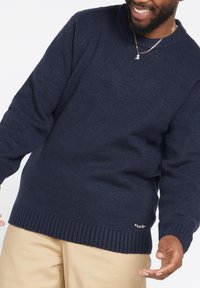 Suéter de punto azul marino con dobladillo y puños acanalados, con cuello redondo y un pequeño parche de logo en la parte inferior izquierda.