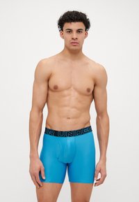 Blaue Boxershorts aus einem glatten, dehnbaren Material mit einem schwarzen elastischen Bund, der weiße Schriftzüge aufweist. Eng anliegendes Design mit einer Vorderpocket.