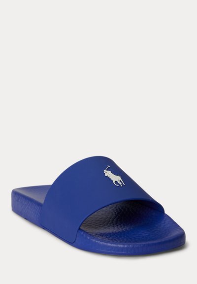 Μπλε σανδάλι με λουράκι και ανάγλυφη ανατομική σόλα, με ασημένιο λογότυπο παίκτη πόλο στο φαρδύ λουράκι, φωτογραφία υπό γωνία πάνω σε απλό ανοιχτό φόντο.