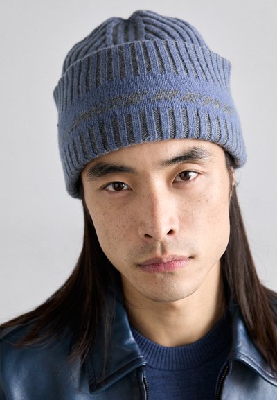 Blue knitted beanie with vertical ribbing and grey accents, featuring a soft texture. The design is snug and fits closely to the head.